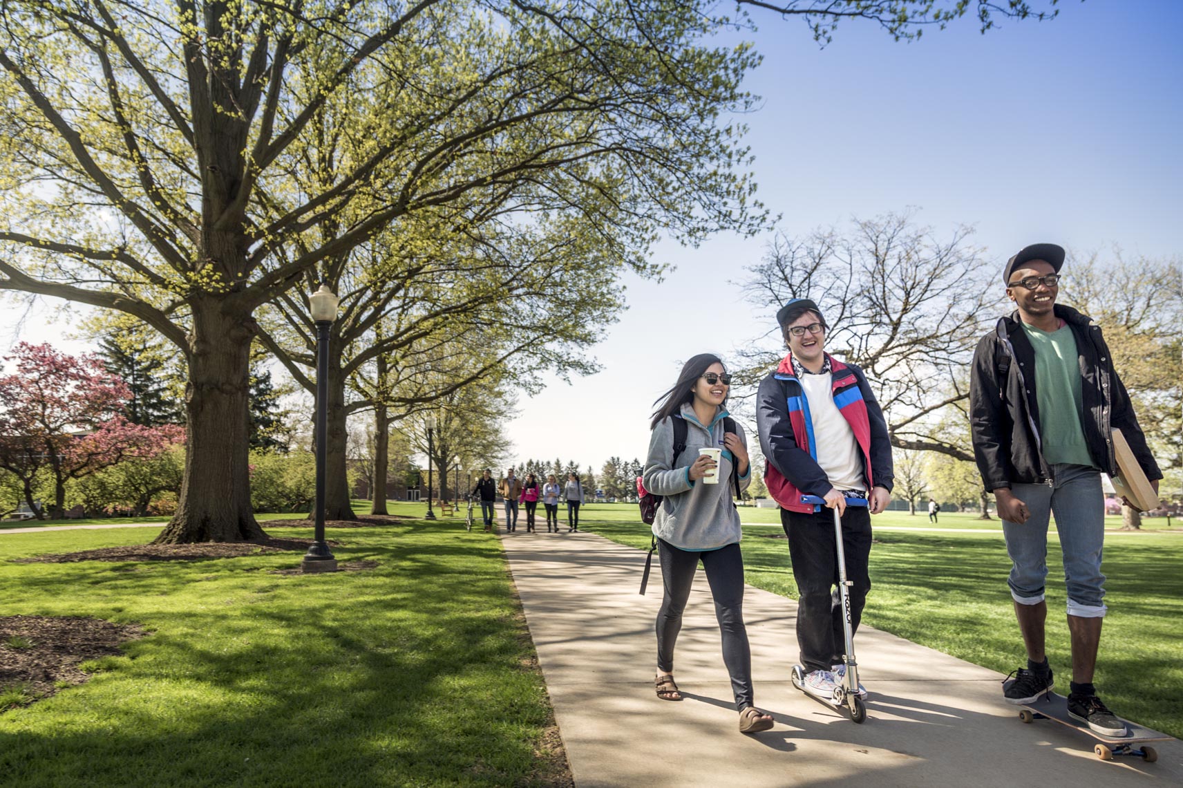 Three college students laugh while walking and scooting on campus in a photo by Ryan Donnell